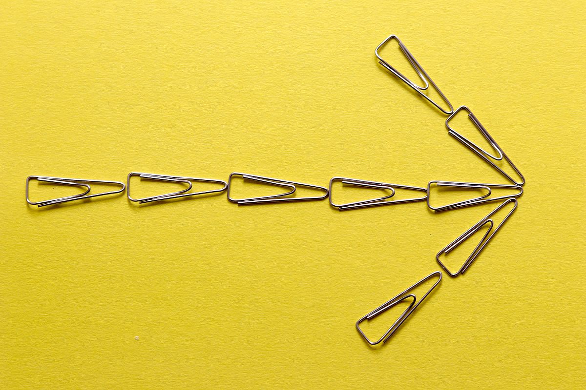 Right-pointing arrow shape made with paper clips on yellow background.