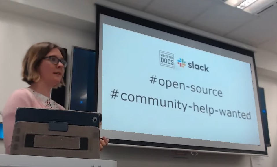 Woman at lectern with projection screen showing hashtags open-source and community-help-wanted