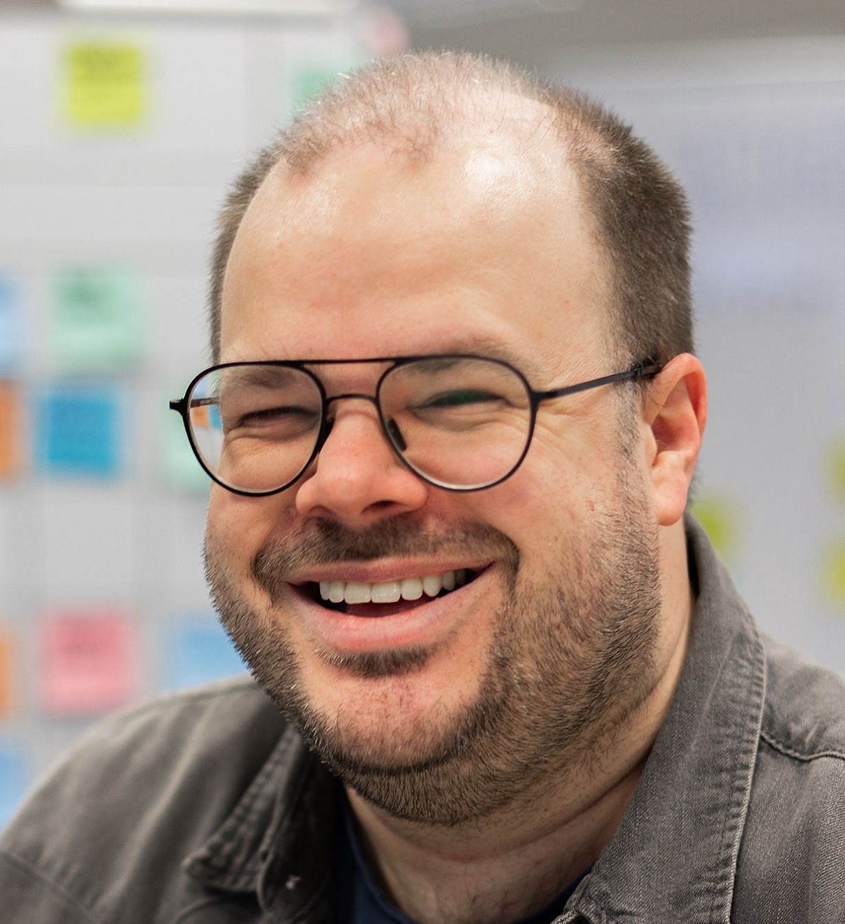 A smiling man with glasses, short hair, and a beard is wearing a dark shirt. He is indoors, with a blurred background featuring colorful sticky notes on a whiteboard.