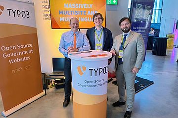 Three men at a booth with an orange TYPO3 design, all dressed business casual.