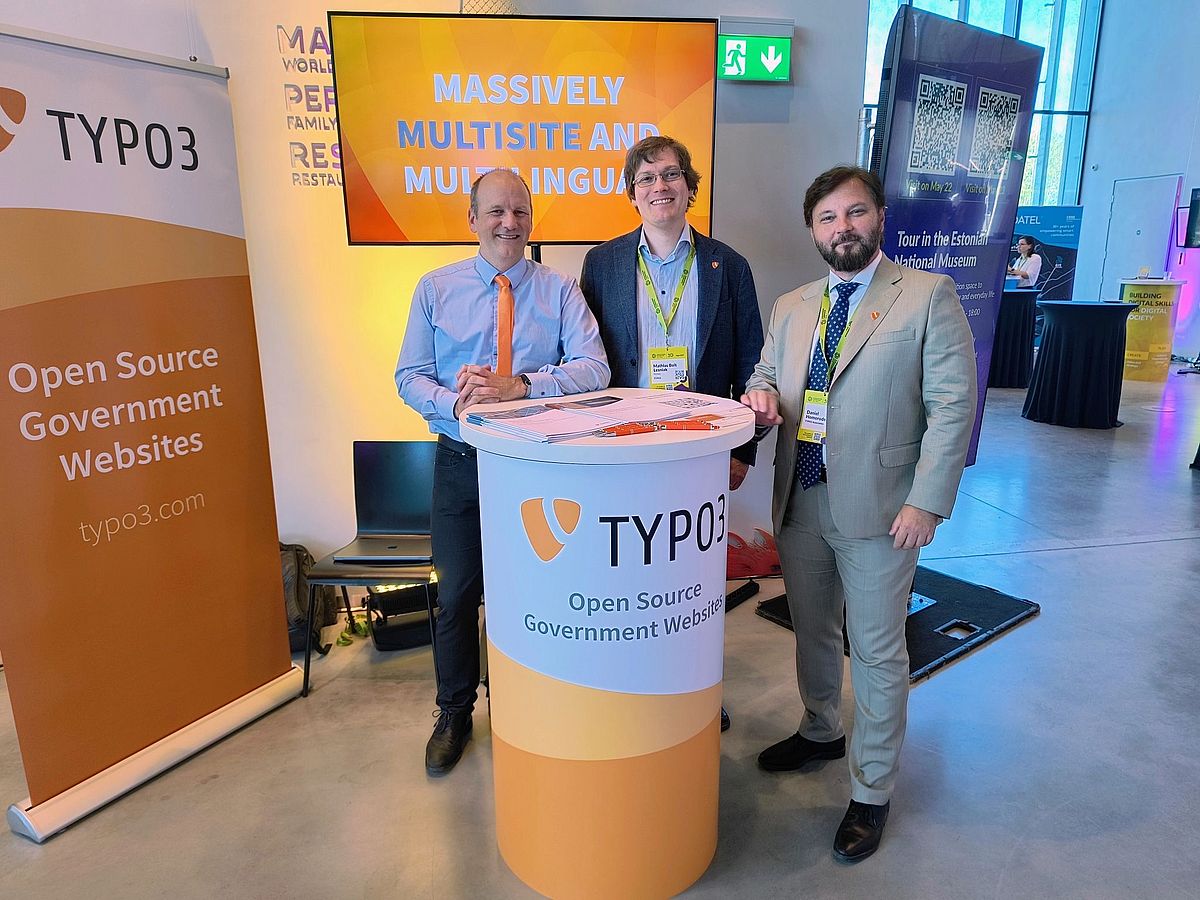 Three men at a booth with an orange TYPO3 design, all dressed business casual.