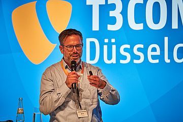 European male in light shirt speaking into a microphone in front of a blue wall saying "T3CON23 Dusseldorf"