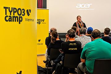 Yellow banners next to a seated crowd watching a woman presenting on stage.