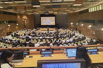 A large conference room filled with people attending a formal meeting. Many sit at desks with laptops. "OSPOs for Good" signs are visible. A speaker is projected on a large screen at the front of the room.
