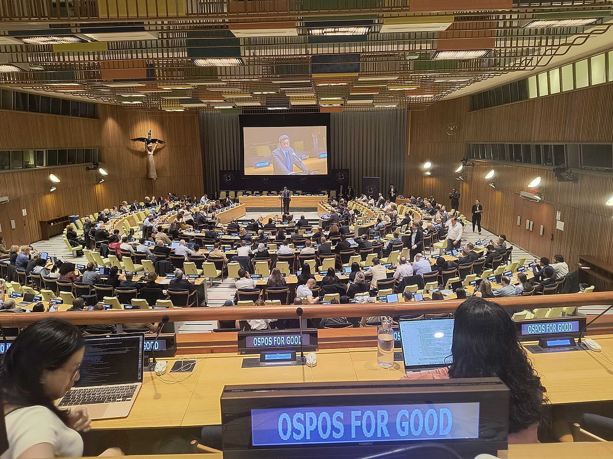 A large conference room filled with people attending a formal meeting. Many sit at desks with laptops. "OSPOs for Good" signs are visible. A speaker is projected on a large screen at the front of the room.