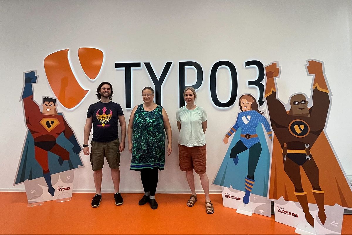 Three people stand smiling in front of a large TYPO3 logo on a wall, flanked by three superhero cardboard cutouts, in a brightly lit room with an orange floor.