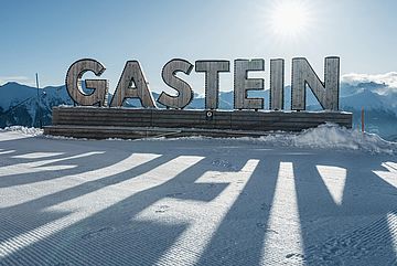 Large wooden letters spelling Gastein stand on a snowy platform with mountains in the background and sunlight casting long shadows on the snow.