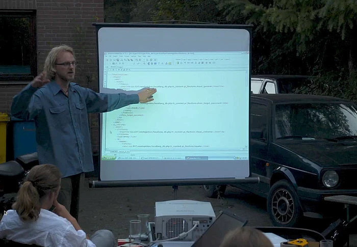 A person stands outdoors pointing at a projection screen displaying code, while others sit and watch; a black car and trees are visible in the background.