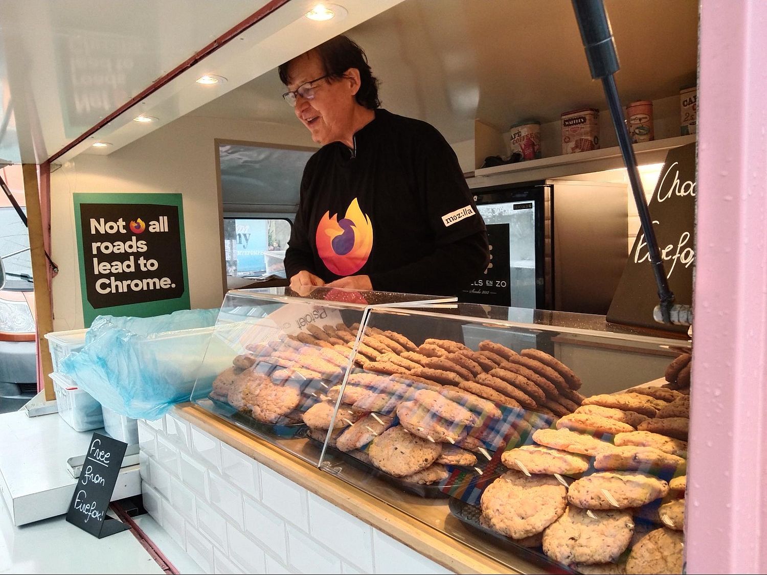 A person stands inside a food truck, wearing a black shirt with a Firefox logo. The truck's counter displays assorted cookies. A sign reads, Not all roads lead to Chrome. The interior has shelves with ingredients and a small refrigerator.