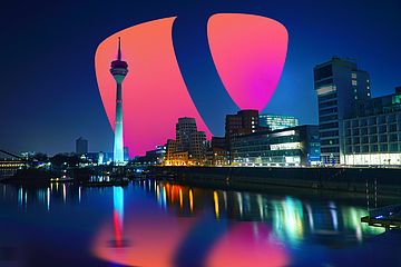 A cityscape at night featuring a tall tower with a glowing red and pink TYPO3 logo in the sky. Buildings are illuminated, reflecting on a calm river in the foreground.