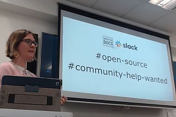 Woman at lectern with projection screen showing hashtags open-source and community-help-wanted