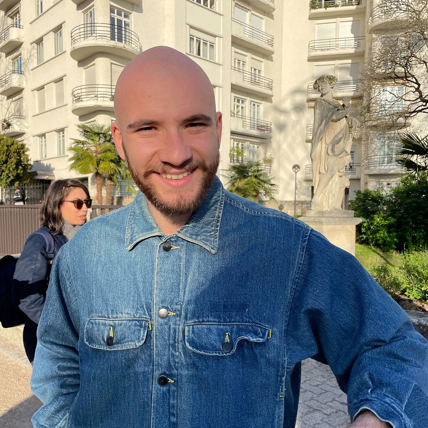 A person with a bald head and beard, wearing a denim jacket, smiles at the camera outdoors. A person in sunglasses and a building with a statue are in the background.