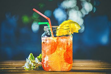 A mason jar filled with a bright orange iced drink with large pieces of fruit, garnished with lemon slices and two straws, sits on a wooden surface with white blossoms beside it and a blurred blue background.