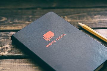 Closed, black notebook with the words "write ideas" in red uppercase sans-serif letters.