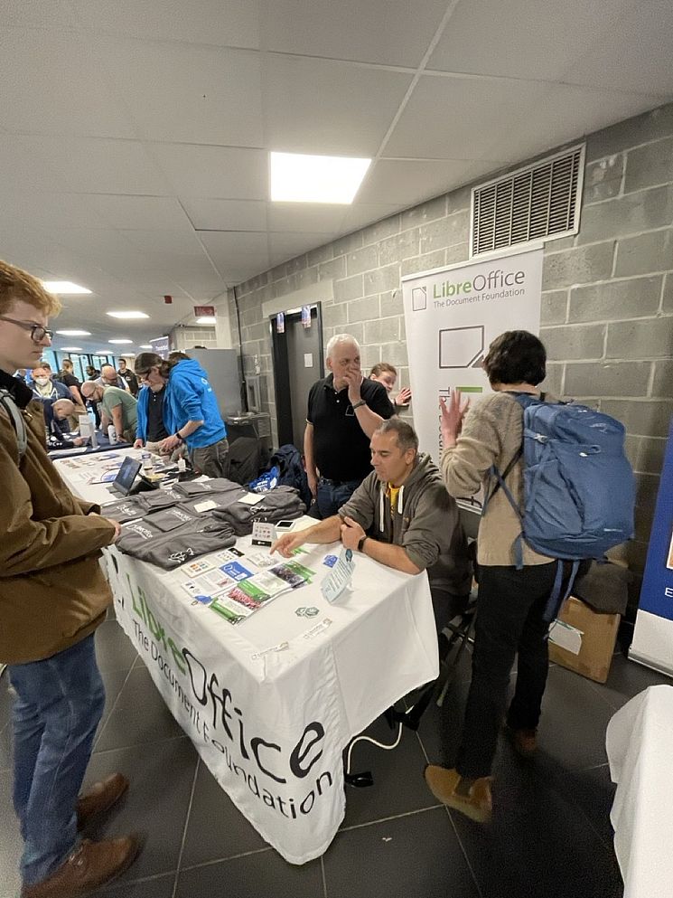 People are gathered at a booth with a LibreOffice banner in a corridor. One person is sitting at the table, while others are standing and interacting. There are pamphlets and merchandise displayed on the table.