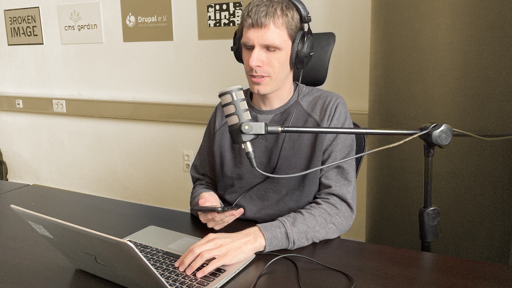Dennis Westphal sitting at a table in front of a microphone with headphones. In his hand he holds a phone, in front of him is a laptop. On the wall behind him, logos of the business units of the company.