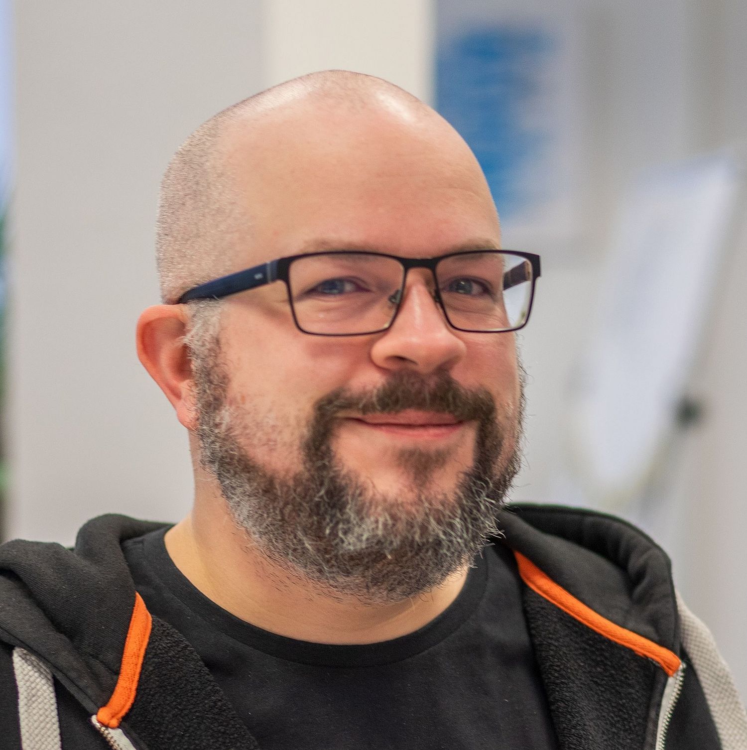 A bearded man with glasses, wearing a black hoodie and a graphic t-shirt, smiles while sitting indoors in a brightly lit office environment.