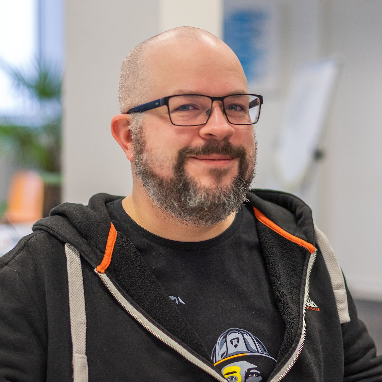 A bearded man with glasses, wearing a black hoodie and a graphic t-shirt, smiles while sitting indoors in a brightly lit office environment.