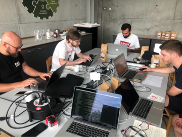 Men working at computers around a table in a concrete-walled meeting room.