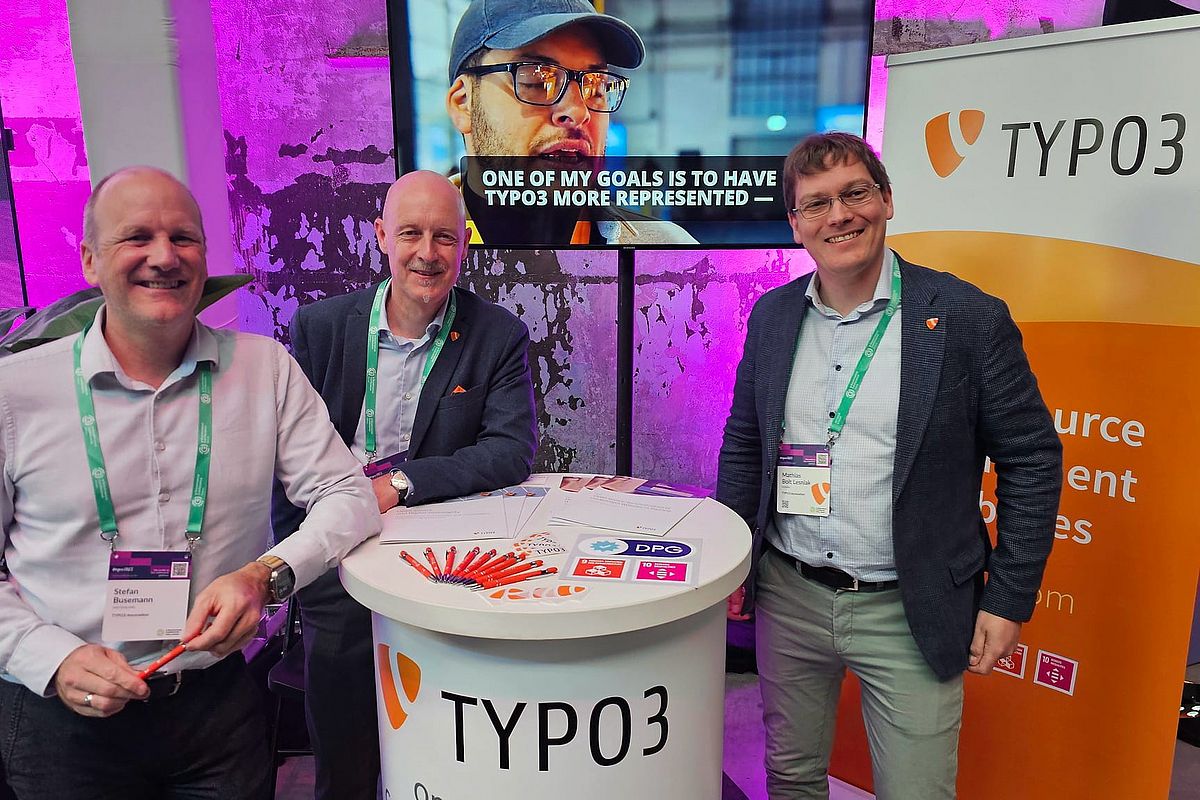 Three smiling men stand at a TYPO3 booth with brochures and pens on the counter. A large screen behind them shows a video with a person and the caption “one of my goals is to have TYPO3 more represented”.