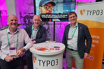 Three smiling men stand at a TYPO3 booth with brochures and pens on the counter. A large screen behind them shows a video with a person and the caption “one of my goals is to have TYPO3 more represented”.