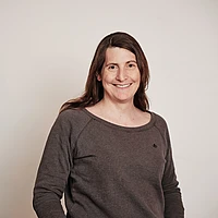 A woman with long brown hair, wearing a grey long-sleeve shirt, stands against a plain light background and smiles at the camera.