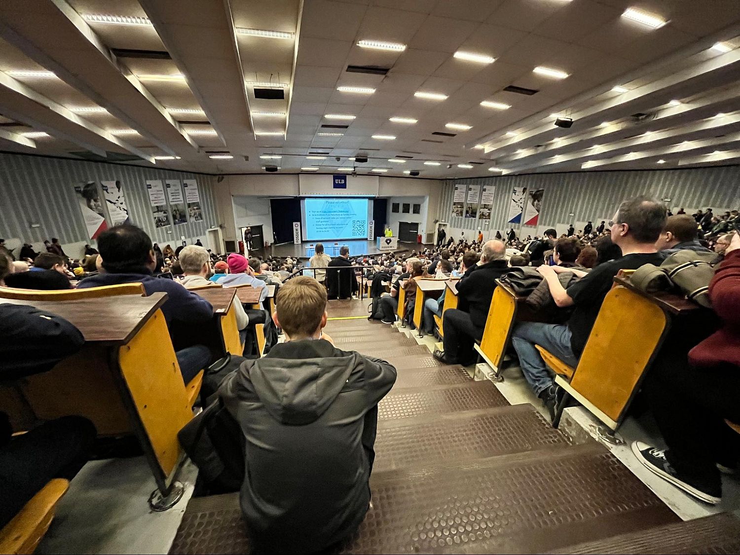 A crowded lecture hall filled with people seated on wooden benches. A large screen at the front displays a presentation slide. The atmosphere is attentive as everyone focuses on the speakers presentation.