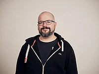 A man with a beard and glasses smiles at the camera. He is wearing a black zip-up hoodie over a dark t-shirt and stands against a plain, light-colored background.