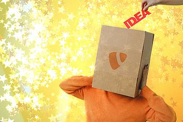 A person in an orange sweater with a cardboard box with the TYPO3 logo over their head stands against a yellow, star-filled background. A hand places the word IDEA into the box.