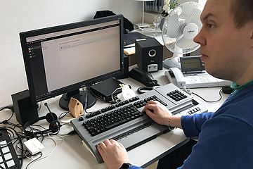 Man sitting in front of a computer screen and Braille display. Screen shows the TYPO3 backend.