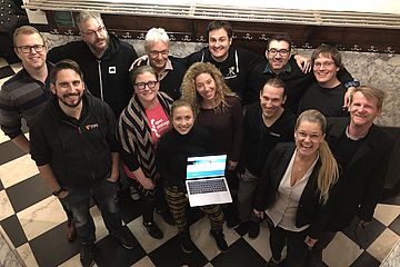 A group of people looking upwards towards the camera, one holding an open computer displaying a web page.