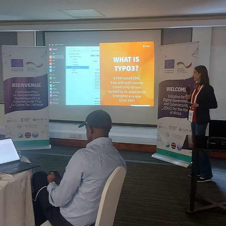 A woman presents a slide titled "What is TYPO3?" to a seated man in a conference room, with two bilingual welcome banners and laptops visible in the background.
