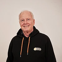 An older man with short white hair smiles at the camera. He is wearing a black hoodie with orange drawstrings and a name badge against a plain, light-colored background.