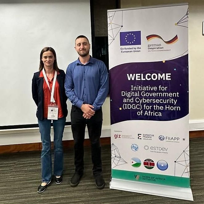 Two people stand side by side in front of a projector screen, between two vertical banners for a digital government and cybersecurity event in Africa, featuring logos of the European Union, German Cooperation, and other partners.
