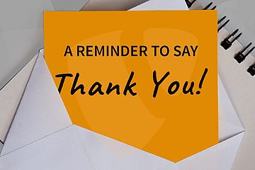 An envelope with an orange card inside reads "A Reminder to Say Thank You!" in black text, placed next to a spiral notebook.