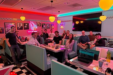 A group of people smile and pose together at a retro-themed diner with neon lights, pastel booths, and vintage decor. Laptops and drinks are on the tables, suggesting a casual meeting or work session.