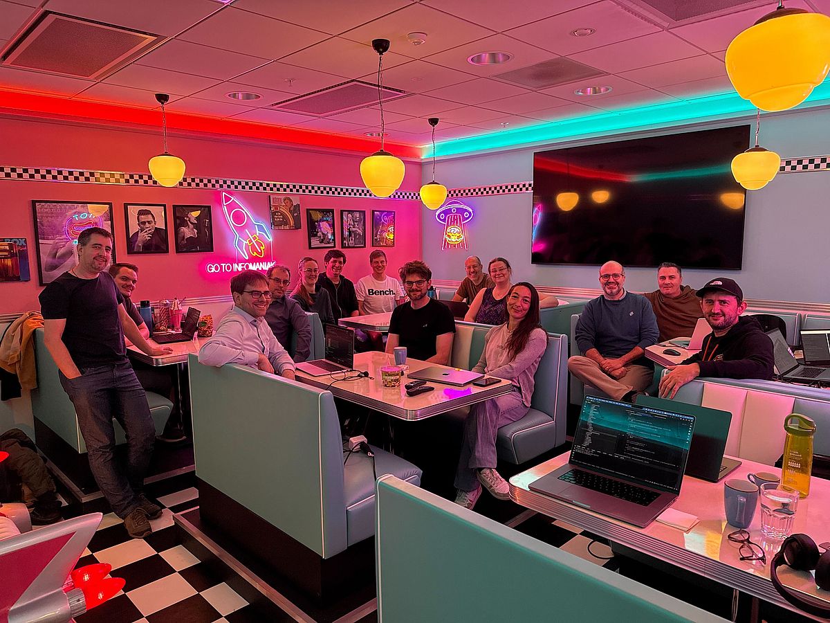 A group of people smile and pose together at a retro-themed diner with neon lights, pastel booths, and vintage decor. Laptops and drinks are on the tables, suggesting a casual meeting or work session.