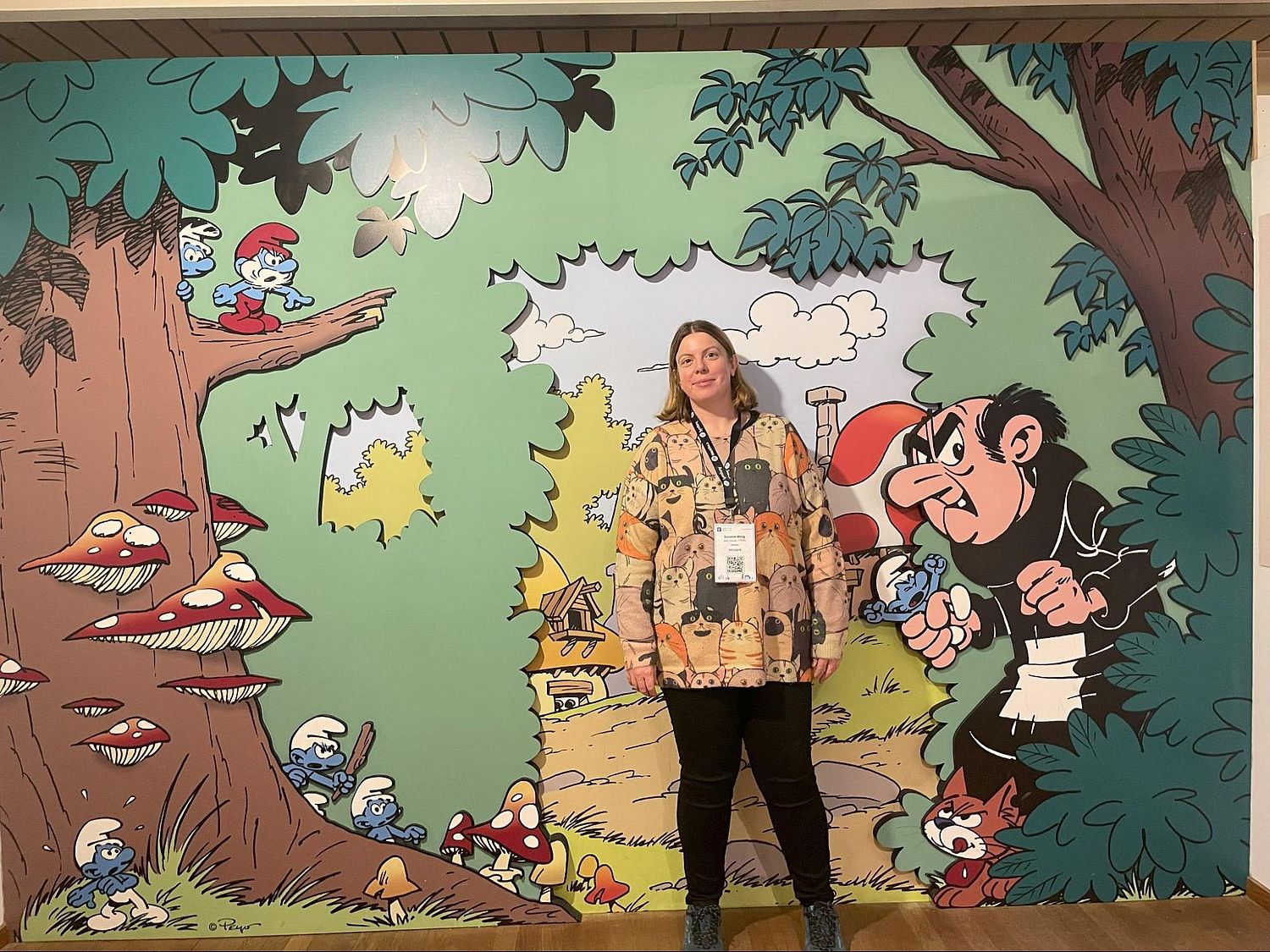 Woman in yellow shirt standing in front of a wall with a Smurf cartoon forest environment with trees and mushrooms.