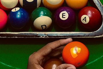 A hand holding an orange billiard ball with the TYPO3 logo in front of a rack of colorful billiard balls spelling "idea". The balls are arranged on a green table, and the rack is slightly worn.