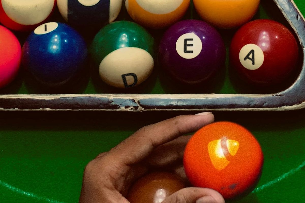 A hand holding an orange billiard ball with the TYPO3 logo in front of a rack of colorful billiard balls spelling "idea". The balls are arranged on a green table, and the rack is slightly worn.