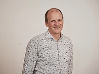 A middle-aged man with a receding hairline smiles warmly while wearing a white shirt with a black floral pattern, standing against a plain, light-colored background.