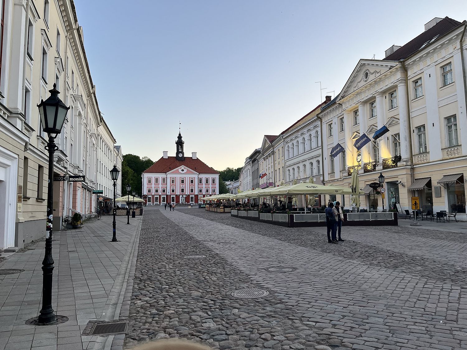 A cobblestone street with outdoor seating on one side and a red and white building with a clock tower at the end, bordered by white historic buildings.