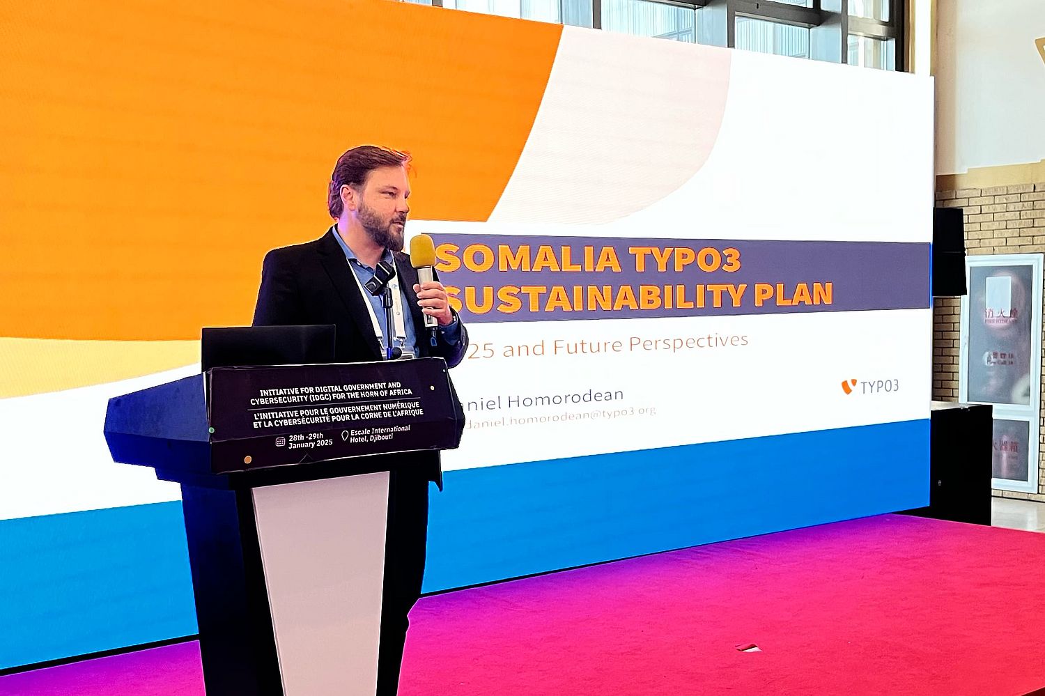 A man in a suit speaks into a microphone at a podium in front of a large screen displaying the text Somalia TYPO3 Sustainability Plan and information about the presentation.