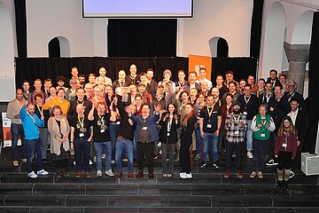 A large group of people stands on a stairway inside a hall, posing for a photo. They are smiling and waving, and some are wearing name badges. The background features a stage with a black curtain and a screen above.