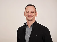 A man with short hair smiles at the camera. He is wearing a dark jacket over a blue patterned button-up shirt, standing in front of a plain, light-colored background.