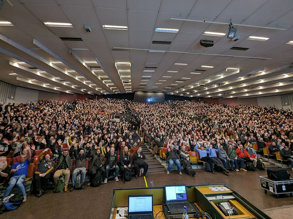 A large crowd fills a lecture hall, with many people raising their hands. The room is packed with attendees seated in rows, and a projector is visible at the rear. The ceiling features multiple rows of lights.