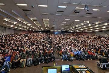 A large crowd fills a lecture hall, with many people raising their hands. The room is packed with attendees seated in rows, and a projector is visible at the rear. The ceiling features multiple rows of lights.