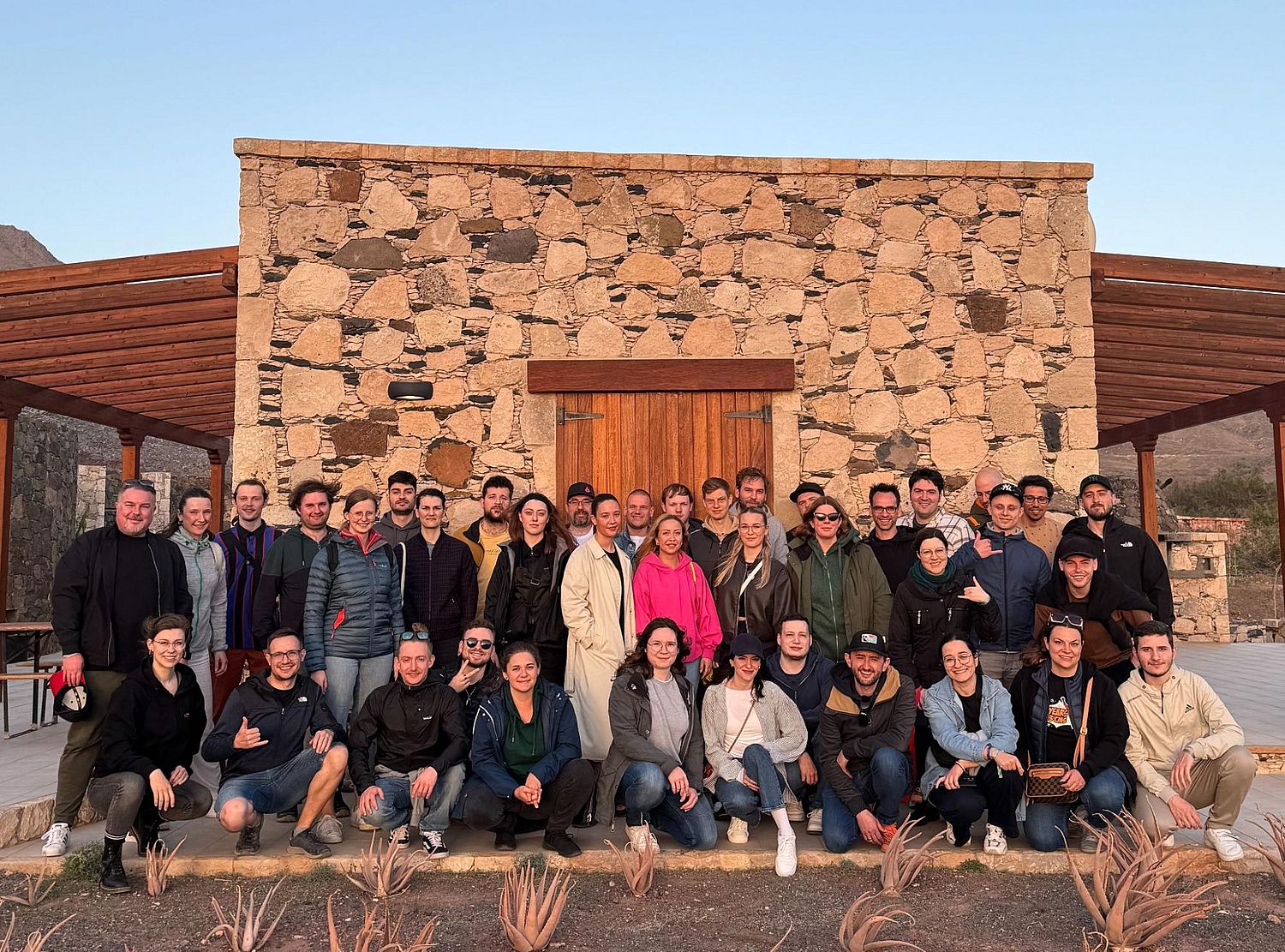 A group of people posing in front of a rustic stone building with wooden elements. Some are sitting, and others are standing.