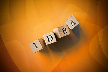 Four wooden blocks spelling IDEA are arranged on an orange abstract background with soft lighting and shadows.