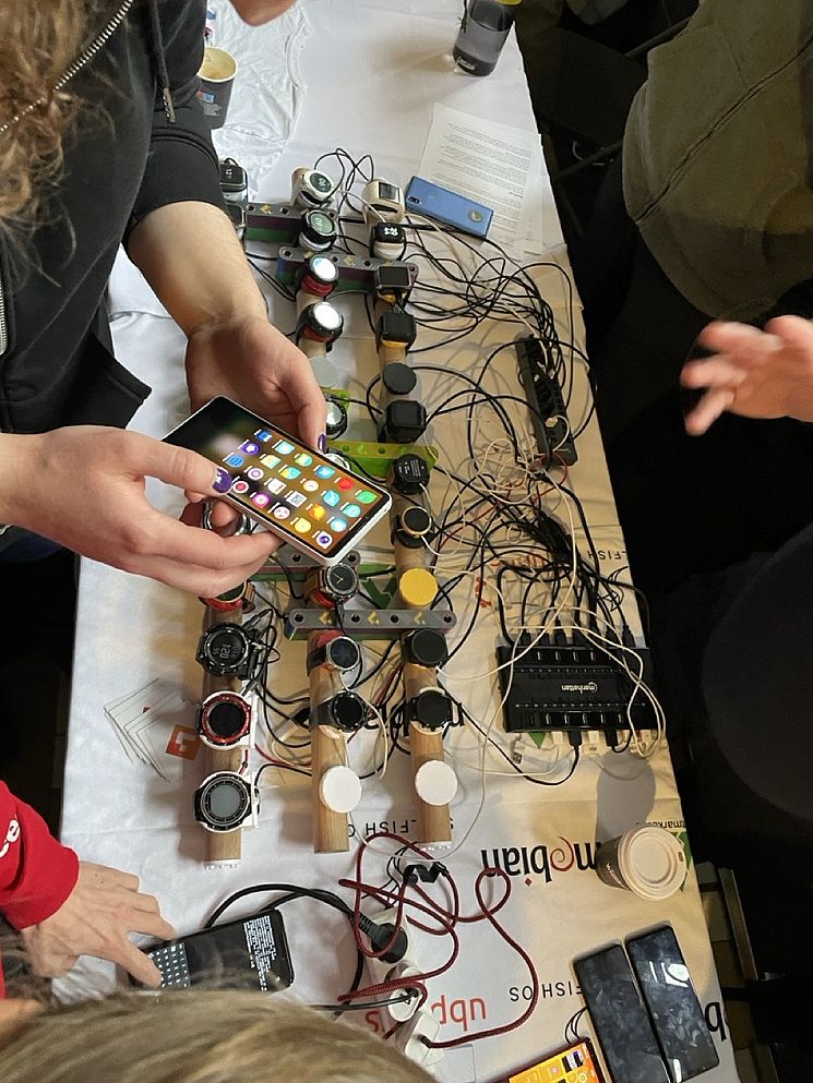 A person holding a smartphone examines various smartwatch models connected by wires on a table. Several individuals are gathered around, engaging with the devices.